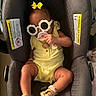 baby, child, car_seat, sunglasses, yellow_outfit, footwear, bracelet, pacifier, hair_bow, infant, accessories, cute, indoors, person, seat, fashion, baby_shoes, smile, relaxing, comfort