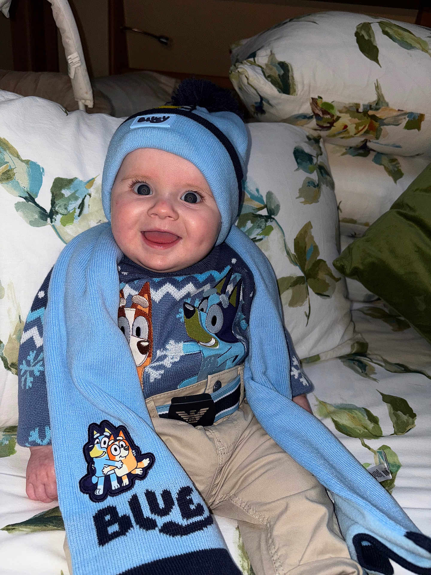 Danny joined the competition — help win amazing prizes! baby, child, smile, hat, scarf, blue, sweater, cartoon, couch, pillow, floral_pattern, indoor, person, clothing, happy, cute, portrait, warm, seated, infant