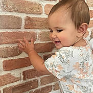 Sara joined the competition — help win amazing prizes! baby, child, toddler, floral_shirt, brick_wall, indoor, smiling, happy, cute, earring, skin, hand, face, head, short_hair, exploring, wall, texture, person, clothing