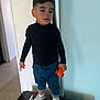 child, toy_car, indoor, black_turtleneck, blue_pants, sneakers, standing, young_boy, casual_clothing, expression, holding, orange_toy, short_hair, light_wall, floor, window_light, home, portrait, person, fashion