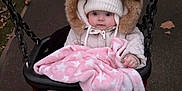 Paisley is registered to the contest to win money with this photo: baby, child, swing, blanket, pink_blanket, fur_hood, white_hat, winter_clothing, outdoor, playground, fall_leaves, pavement, cold_weather, cute, infant, seated, person, face, footwear, cozy