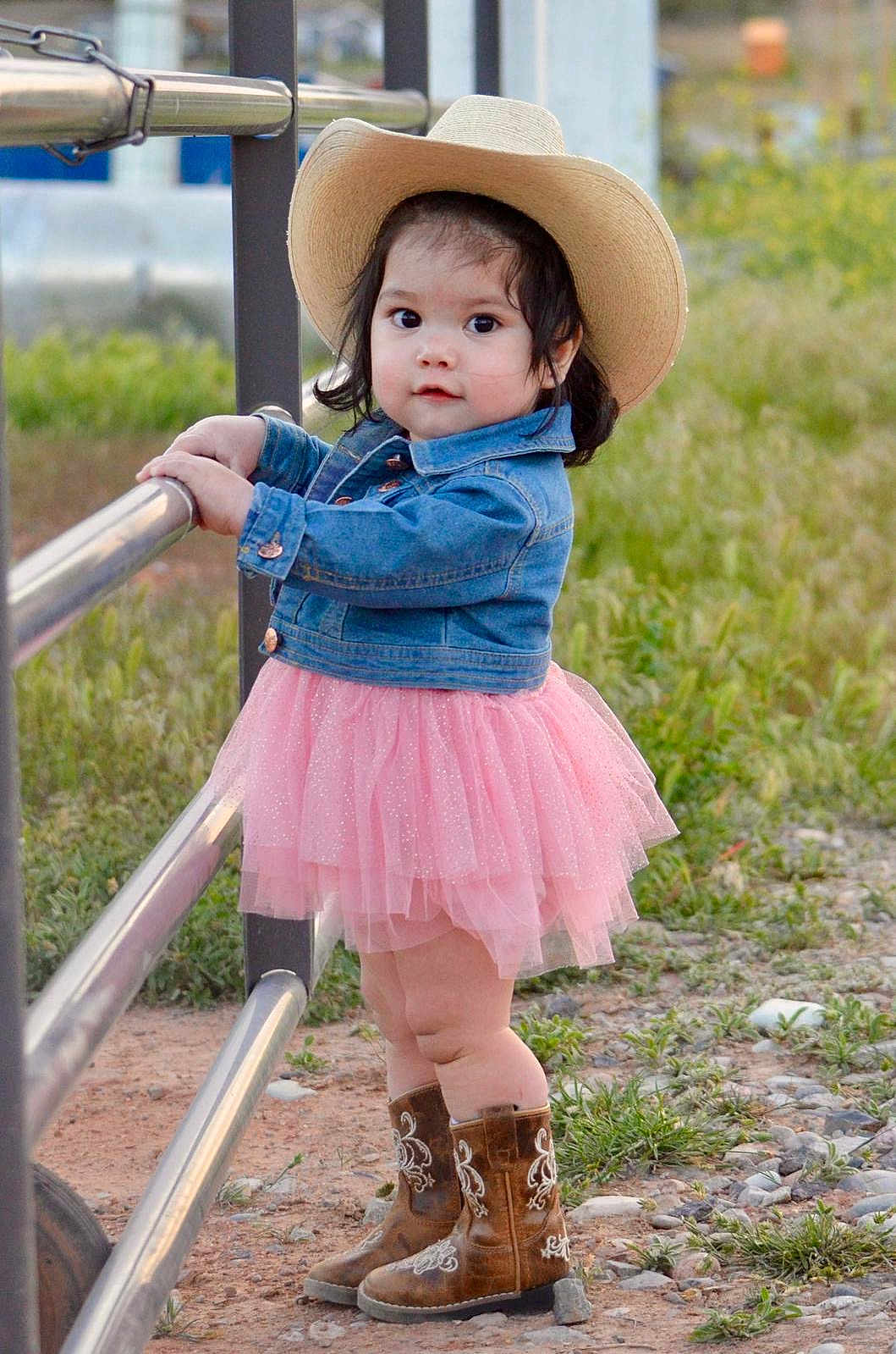 Arianna is registered to the contest to win money with this photo: toddler, child, cowboy_hat, denim_jacket, pink_tutu, cowboy_boots, outdoor, fence, grass, dirt, curious, standing, young_child, nature, casual_clothing, portrait, cute, baby, person, daylight