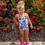 child, girl, swimsuit, pink_crocs, flower_pattern, standing, outdoor, plants, greenery, red_flowers, sidewalk, hair_bun, summer, confident, nature, garden, young_child, casual, daylight, portrait
