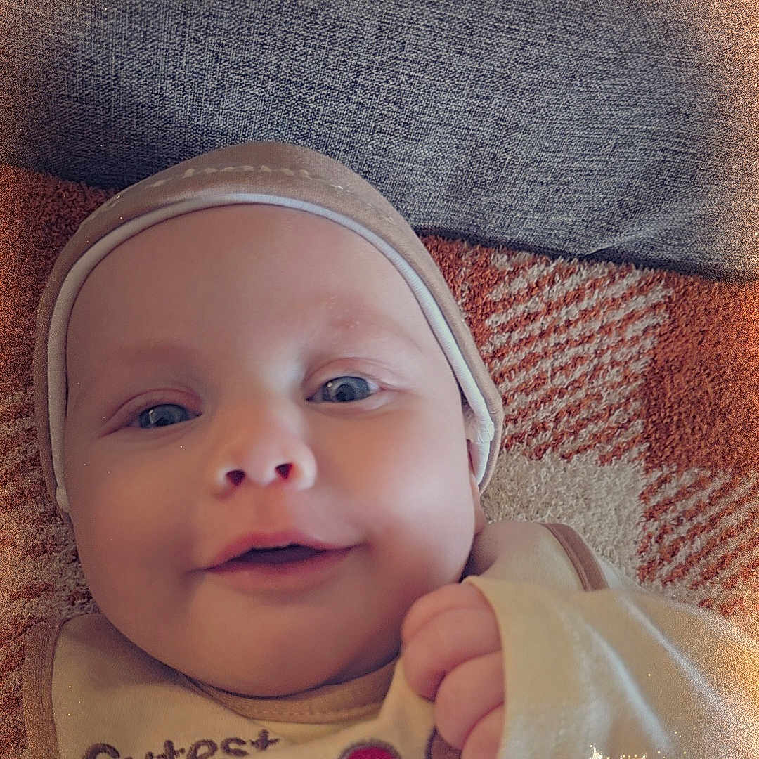 Jensen joined the competition — help win amazing prizes! baby, blanket, child, closeup, clothing, cute, face, happy, hat, headwear, indoors, infant, lying_down, newborn, person, portrait, skin, smile, soft_texture, warm_colors
