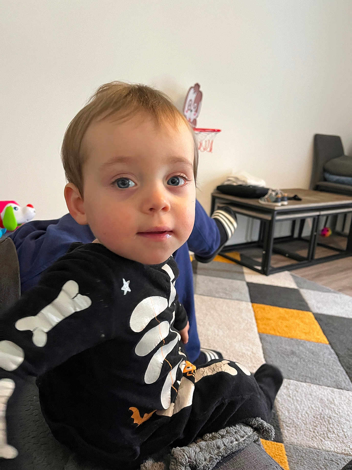 Noah participe au concours pour gagner de l'argent avec cette photo : toddler, child, boy, indoor, couch, skeleton_outfit, carpet, basketball_hoop, toy, face, blue_eyes, hair, clothing, furniture, floor, person, home, casual, playful, portrait