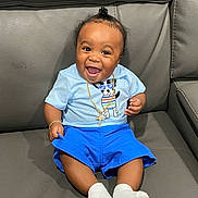Tru is registered to the contest to win money with this photo: baby, child, smiling, happy, blue_shorts, light_blue_shirt, dog_graphic, gold_chain, bracelet, socks, ponytail, couch, leather, indoor, portrait, cute, sitting, person, infant, joyful