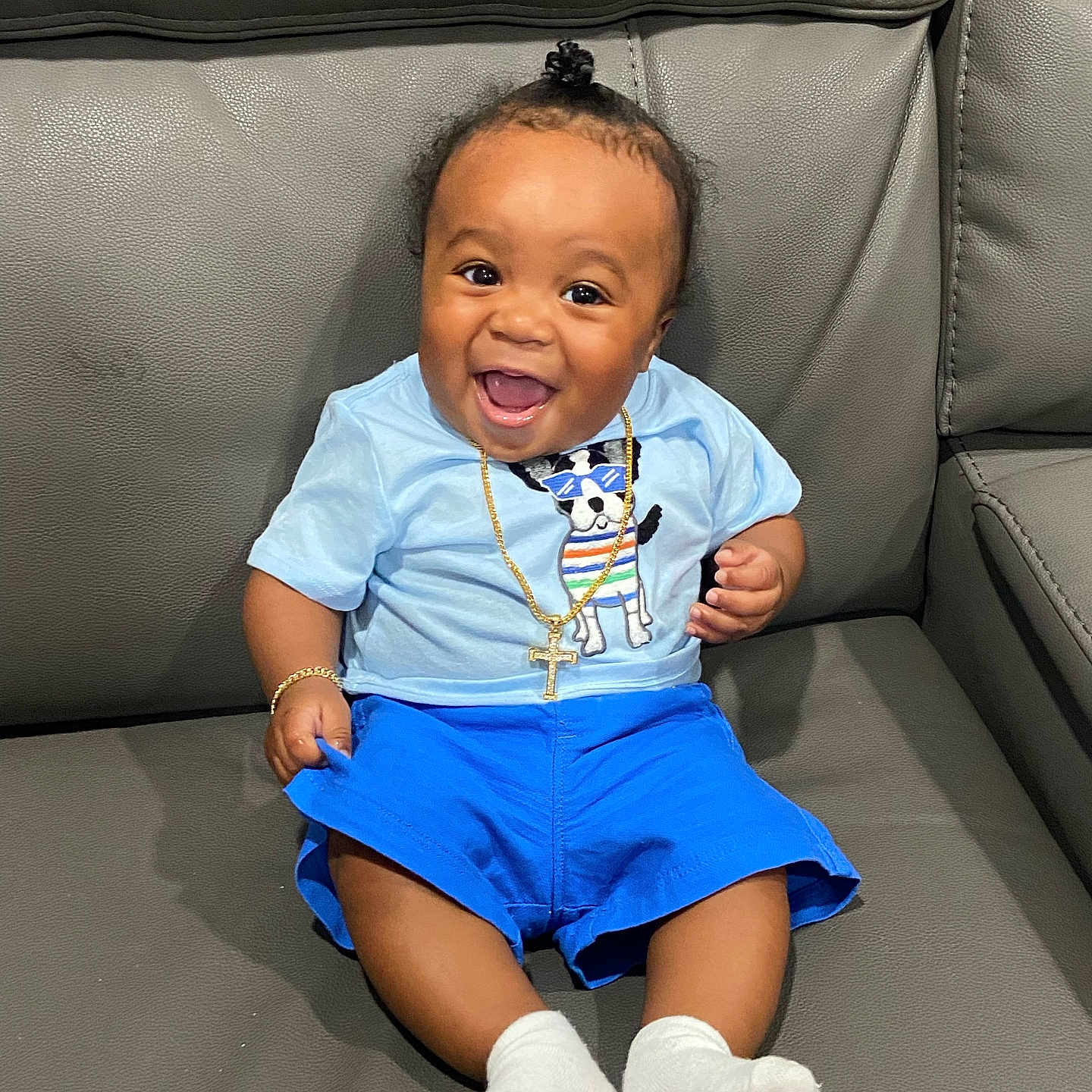 Tru is registered to the contest to win money with this photo: baby, child, smiling, happy, blue_shorts, light_blue_shirt, dog_graphic, gold_chain, bracelet, socks, ponytail, couch, leather, indoor, portrait, cute, sitting, person, infant, joyful