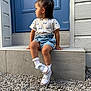 toddler, child, sitting, blue_door, stone_steps, denim_shorts, white_sneakers, white_socks, gravel, outdoor, curious, side_profile, short_hair, casual_clothing, daylight, young_child, front_entrance, building_wall, footwear, portrait