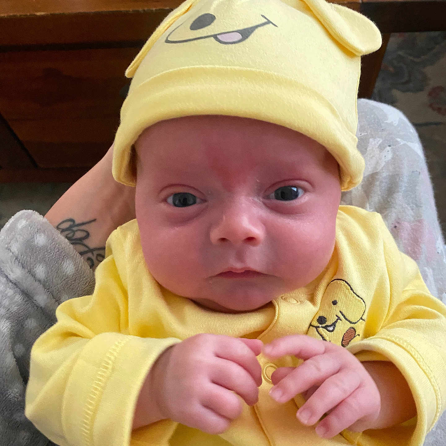 Luca joined the competition — help win amazing prizes! adult_hand, baby, baby_clothes, blue_eyes, close_up, cozy, cute, face, hands, indoor, infant, newborn, person, portrait, soft_robe, table, tattoo, wooden_furniture, yellow_hat, yellow_outfit