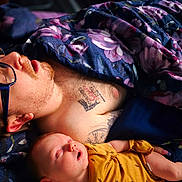 Aiden joined the competition — help win amazing prizes! man, baby, sleeping, glasses, tattoos, blanket, floral_pattern, yellow_clothing, closeup, face, rest, nap, indoor, family, bonding, relaxation, portrait, skin, peaceful, comfort