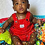 Zakai is registered to the contest to win money with this photo: baby, child, infant, red_overalls, confetti, colorful, quilt, sitting, curious, expression, bright, indoors, portrait, cute, hands, feet, face, clothing, playful, happy