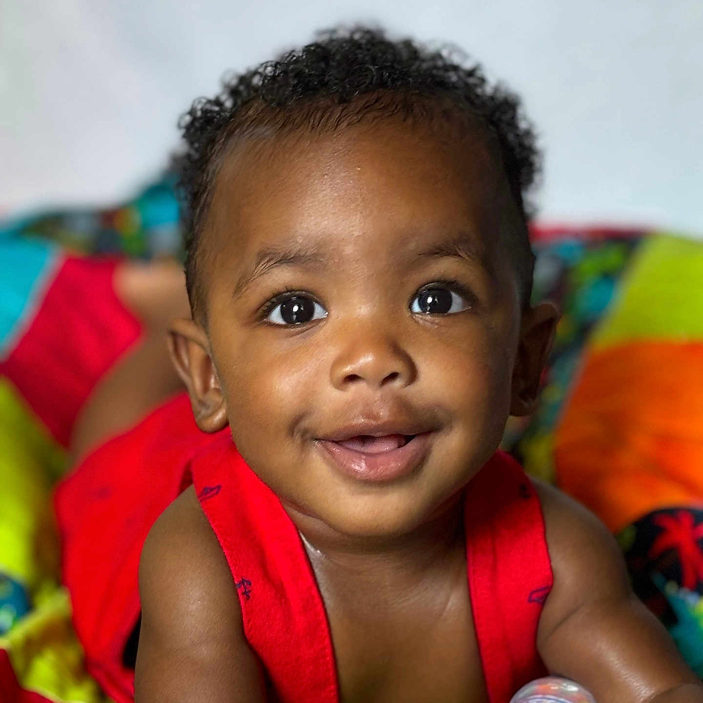 Zakai is registered to the contest to win money with this photo: baby, child, closeup, colorful_blanket, curly_hair, cute, expression, eyes, face, happy, indoors, infant, lying_down, person, portrait, red_clothing, skin, smiling, toddler, young_child
