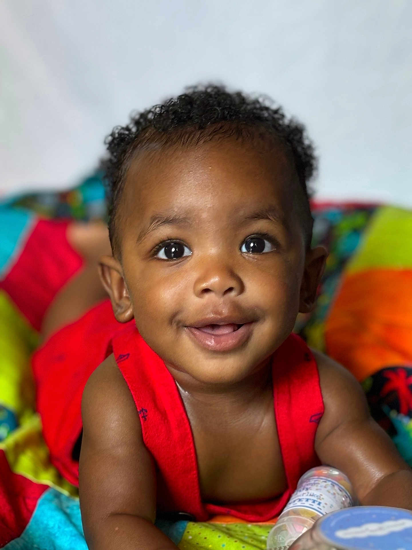 Zakai is registered to the contest to win money with this photo: toddler, child, smiling, face, eyes, curly_hair, red_clothing, colorful_blanket, portrait, baby, cute, happy, skin, infant, lying_down, closeup, expression, indoors, person, young_child