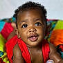 baby, child, infant, red_clothing, colorful_quilt, bottle, smiling, curious, lying_down, close_up, face, bright_eyes, indoor, soft_light, happy, cute, skin, person, portrait, young_child