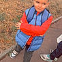 child, boy, standing, outdoor, puffer_vest, red_sweatshirt, black_pants, sneakers, sidewalk, grass, street, people, backpack, crossed_arms, casual_clothing, short_hair, daylight, urban, confident, fashion