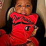 baby, infant, child, face, clothing, red_outfit, black_sleeves, pacifier, pacifier_clip, car_seat, person, portrait, cute, young_child, indoors, seated, expression, wide_eyes, hand, soft_lighting