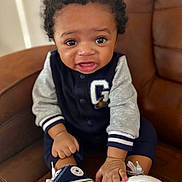 Jahloni Brown is registered to the contest to win money with this photo: baby, child, curly_hair, sneakers, navy_jacket, gray_sleeves, brown_chair, leather_chair, indoor, cute, portrait, small_hands, polka_dots, footwear, sitting, looking_at_camera, expression, infant, clothing, person