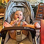 baby, stroller, snack, indoor, cafe, child, food, happy, feet, seatbelt, diaper, light, wall_art, table, chair, person, wood_floor, casual, playful, smiling