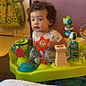 toddler, child, baby, sitting, portrait, curly_hair, facial_expression, indoors, activity_center, baby_toys, colorful, fish_toy, playtime, infant, shirt, home, furniture, carpet, bucket, wooden_door
