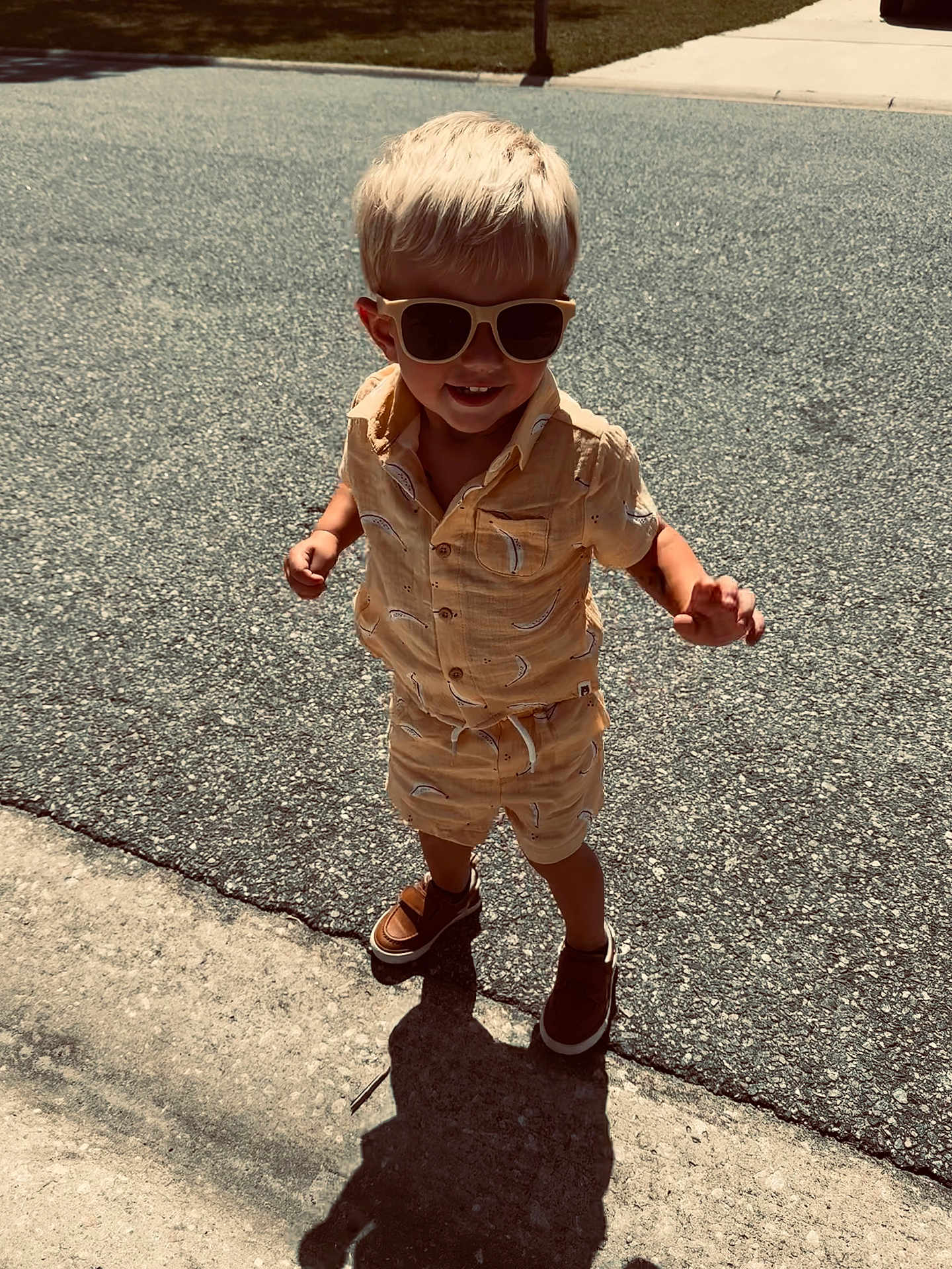 Weston joined the competition — help win amazing prizes! toddler, child, blonde_hair, sunglasses, shorts, shirt, banana_print, shoes, sidewalk, street, shadow, sunny, outdoor, smiling, walking, summer, casual_clothing, happy, person, daytime