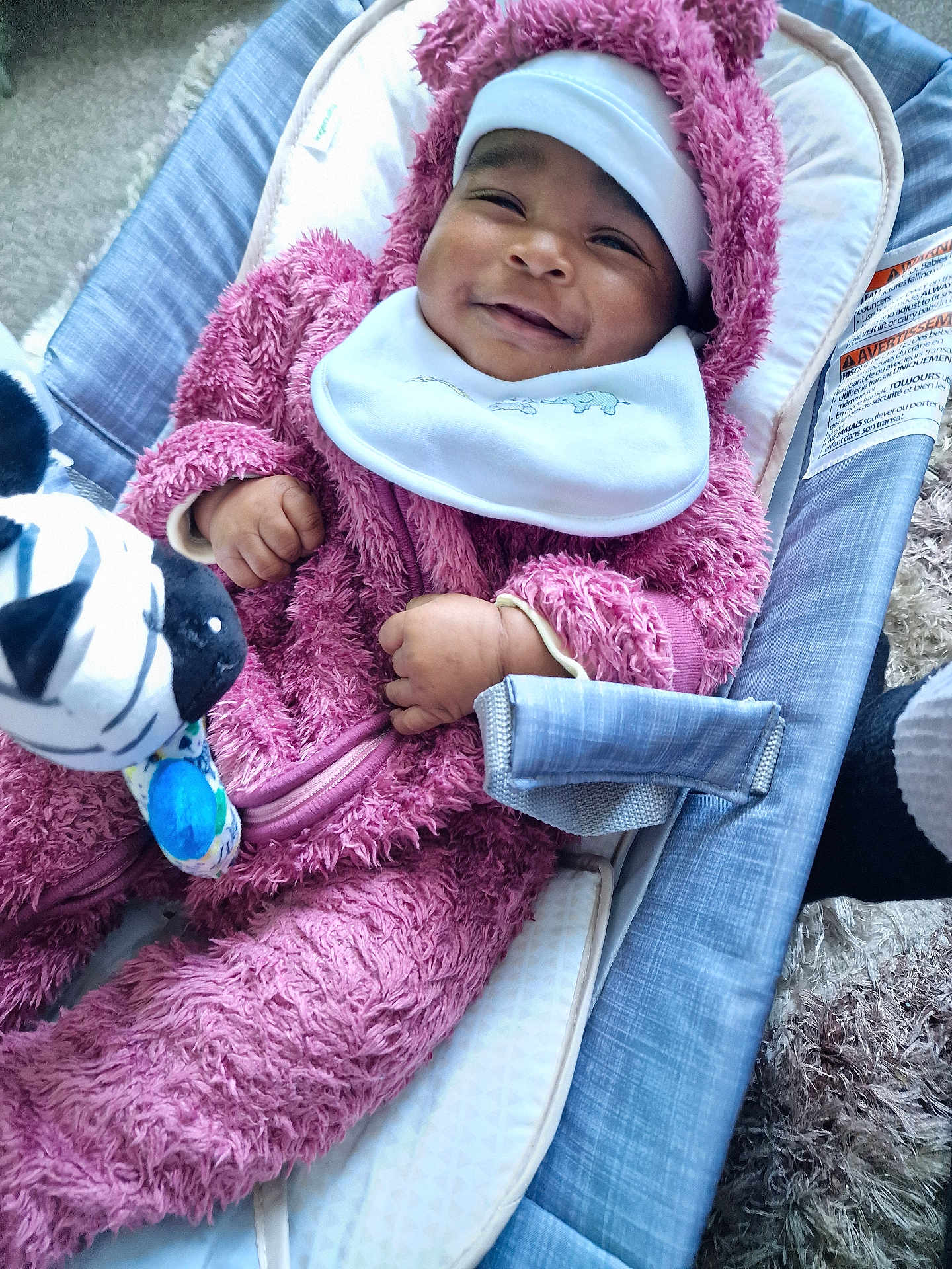 Talia-Jane joined the competition — help win amazing prizes! baby, infant, smile, pink_onesie, fluffy_clothing, baby_seat, toy, zebra_toy, pacifier, white_bib, headwear, happy, cozy, indoor, carpet, cushion, child, person, cute, relaxed