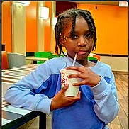 Syxia is registered to the contest to win money with this photo: blue_sweater, braids, candid, casual, child, drink, floor, green_chair, hairpin, indoor, lighting, orange_wall, person, portrait, relaxed, seated, straw, striped_table, table, young