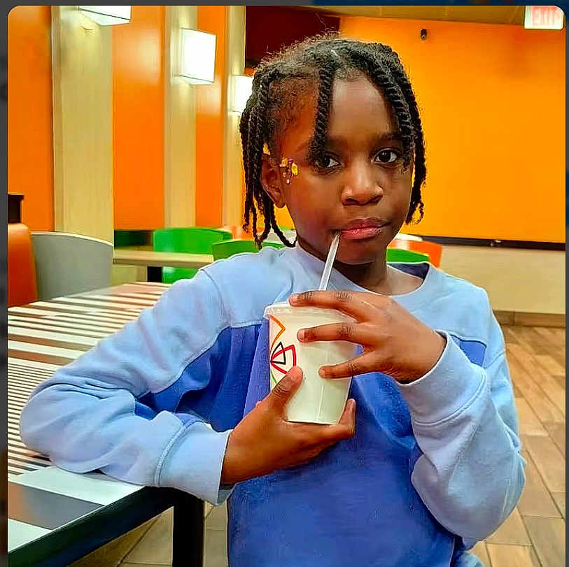 Syxia is registered to the contest to win money with this photo: blue_sweater, braids, candid, casual, child, drink, floor, green_chair, hairpin, indoor, lighting, orange_wall, person, portrait, relaxed, seated, straw, striped_table, table, young