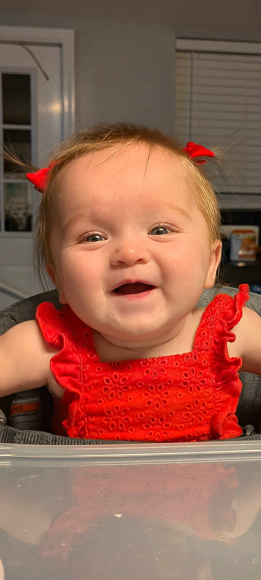 Oakley is registered to the contest to win money with this photo: baby, child, smiling, happy, red_dress, hair_bows, high_chair, reflection, indoor, face, cute, infant, person, portrait, eyes, cheeks, clothing, window_blinds, door, home