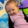 Oakley is registered to the contest to win money with this photo: baby, child, smile, purple_dress, plush_toys, blue_pillow, green_pillow, black_sleeves, face, person, infant, cute, indoors, portrait, happy, young_child, soft_toys, cushion, baby_girl, play_area