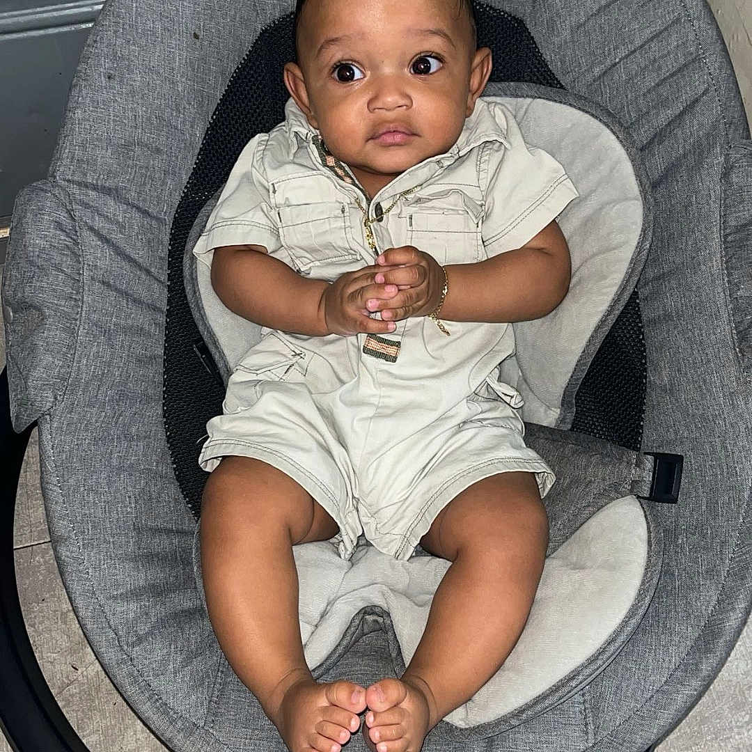 Denzel joined the competition — help win amazing prizes! baby, baby_rocker, baby_seat, black_hair, child, clothing, curious_expression, cute, feet, floor, floor_tile, gray_cushion, hands, indoor, infant, person, romper, seat, sitting, wide_eyes