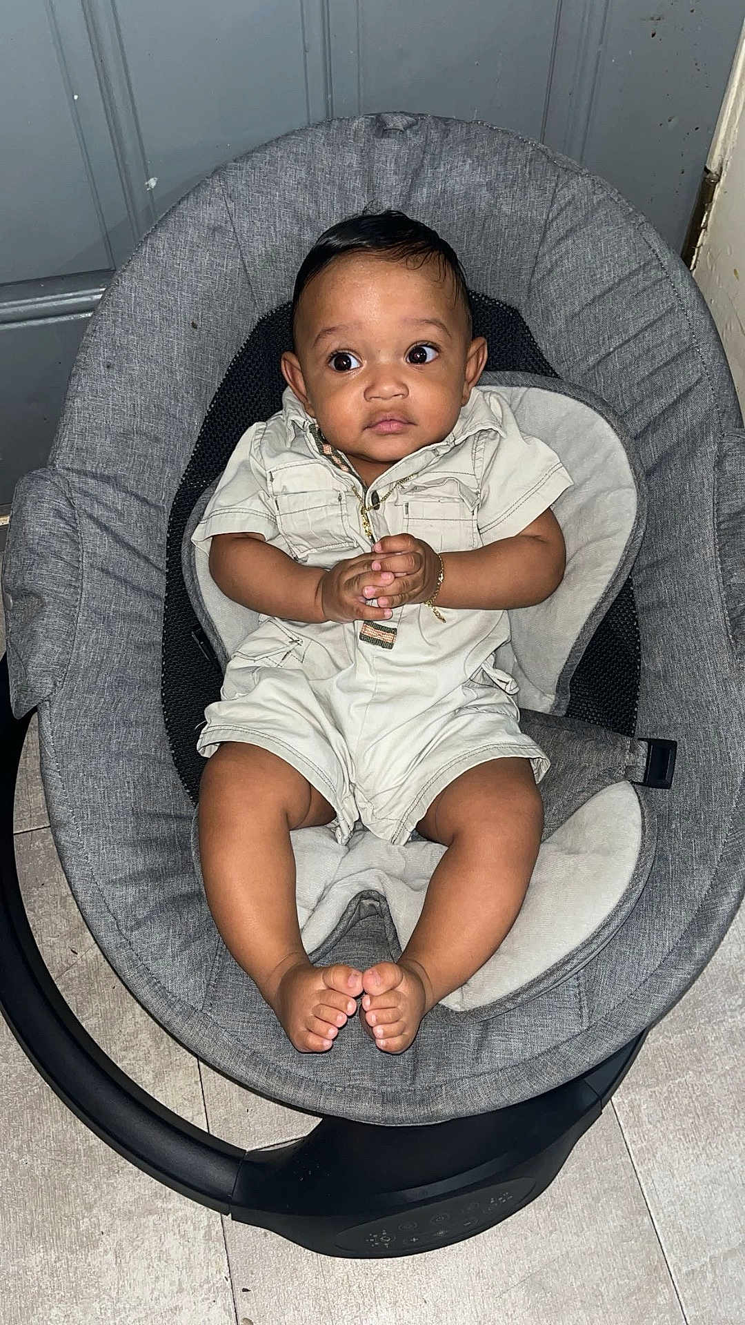 Denzel joined the competition — help win amazing prizes! baby, infant, child, baby_rocker, gray_cushion, romper, clothing, hands, feet, floor, indoor, curious_expression, wide_eyes, black_hair, person, sitting, seat, baby_seat, floor_tile, cute