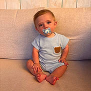 Kyro is registered to the contest to win money with this photo: baby, child, pacifier, blue_outfit, teddy_bear_patch, sitting, couch, beige, wooden_background, indoors, cute, portrait, infant, small_hands, barefoot, expression, soft_lighting, casual_clothing, looking_at_camera, young