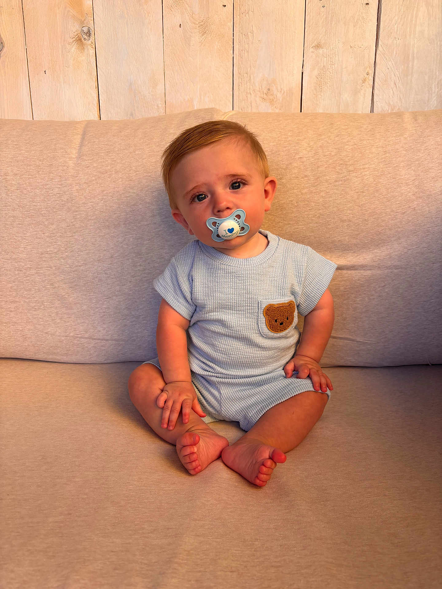 Kyro is registered to the contest to win money with this photo: baby, child, pacifier, blue_outfit, teddy_bear_patch, sitting, couch, beige, wooden_background, indoors, cute, portrait, infant, small_hands, barefoot, expression, soft_lighting, casual_clothing, looking_at_camera, young