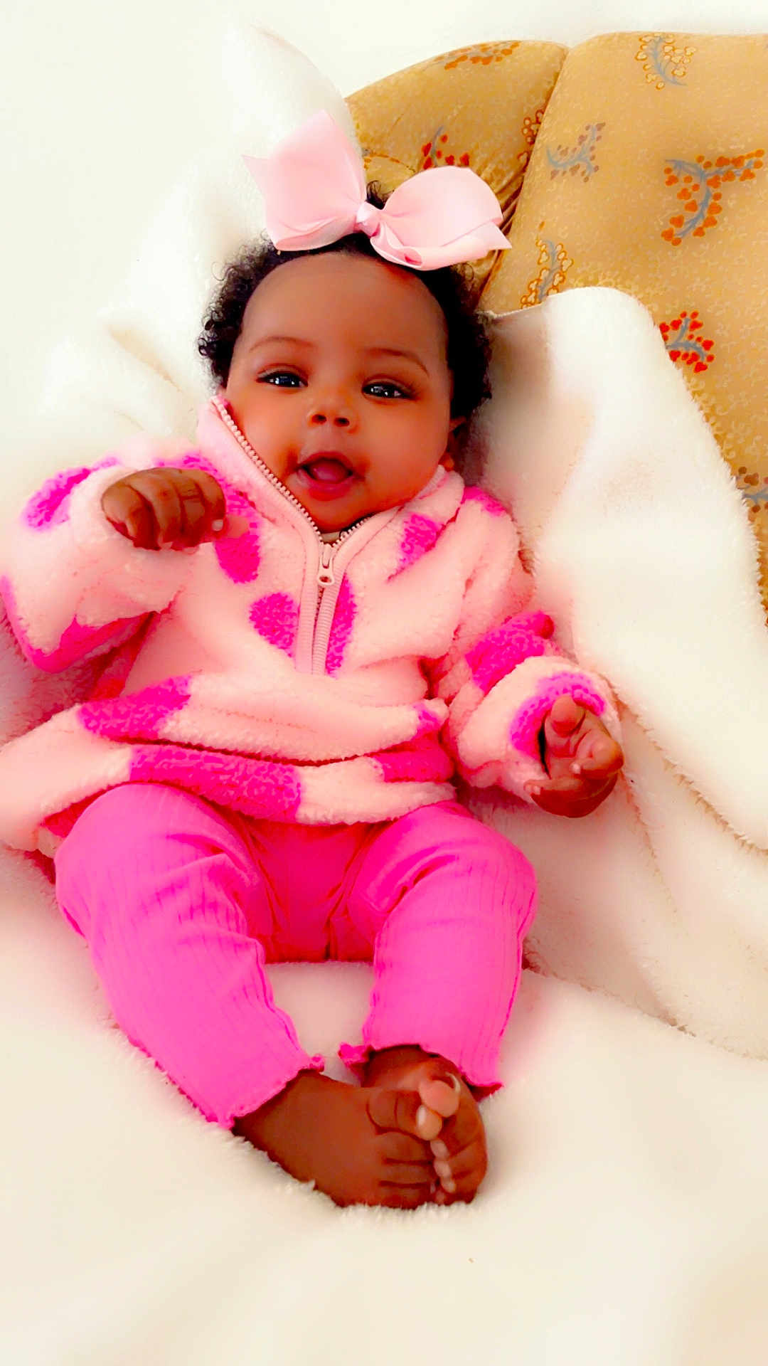 Anaria joined the competition — help win amazing prizes! baby, infant, pink_clothing, bow, curly_hair, blanket, soft_texture, happy_expression, indoors, couch, child, cute, portrait, smiling, fuzzy_jacket, feet, hands, relaxed, cozy, young_child