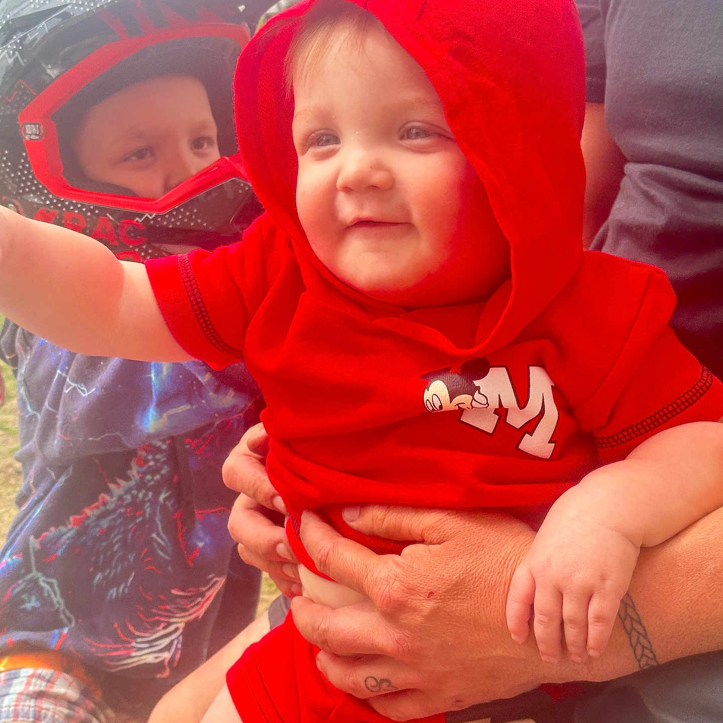 Corbin joined the competition — help win amazing prizes! child, toddler, baby, helmet, red_clothing, mickey_mouse, adult_hand, outdoor, smile, dinosaur_tshirt, person, holding, hoodie, shorts, face, headwear, tattoo, playful, curious, casual
