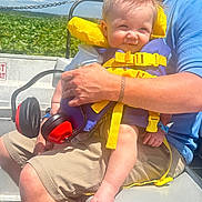 Corbin is registered to the contest to win money with this photo: baby, child, life_jacket, earmuffs, adult, boat, water_plants, sky, blue_shirt, shorts, summer, outdoor, sunlight, person, nature, greenery, smile, hair_spikes, safety, holding