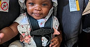 Justice is registered to the contest to win money with this photo: baby, child, car_seat, smiling, hat, dress, bottle, safety_harness, infant, cute, happy, person, seat, seatbelt, warning_label, indoors, skin, hand, clothing, portrait