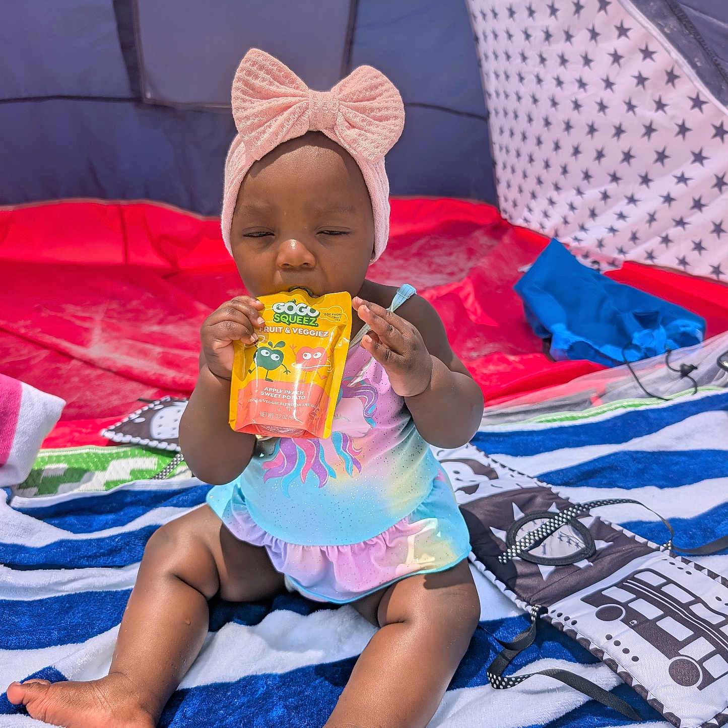 Justice joined the competition — help win amazing prizes! baby, child, toddler, swimsuit, headband, bow, snack_pouch, tent, striped_towel, beach, sunlight, sitting, outdoor, colorful, foot, hand, face, clothing, play, summer