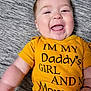 baby, child, smiling, tongue_out, yellow_shirt, text_on_clothing, lying_down, close_up, cute, happy, infant, face, skin, earrings, red_bracelet, soft_surface, texture, casual, indoor, portrait
