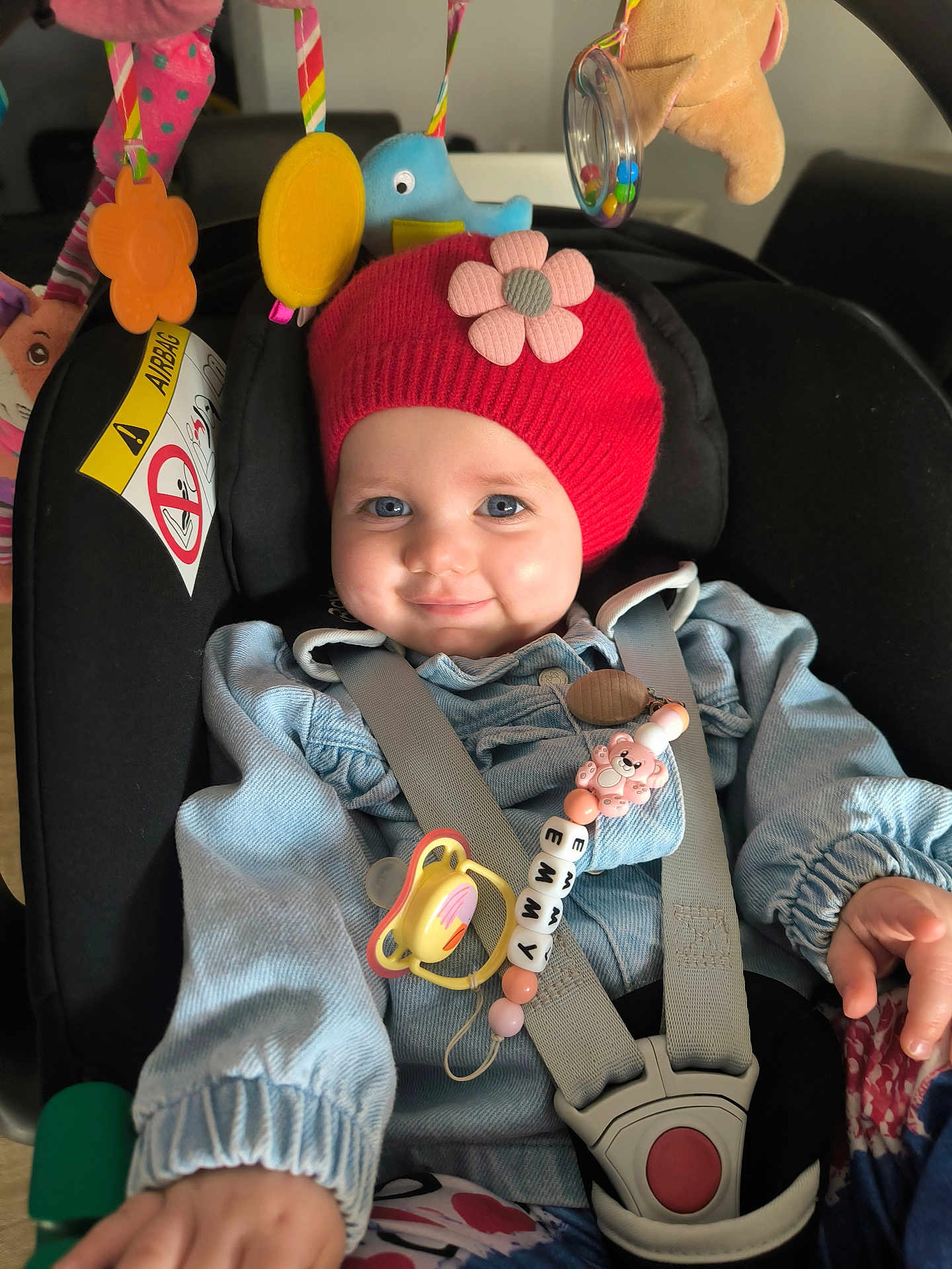 Emmy participe au concours pour gagner de l'argent avec cette photo : baby, infant, child, car_seat, pacifier, beanie, red_hat, flower_decor, toy_mobile, plush_toy, seatbelt, buckle, smiling, blue_eyes, denim_jacket, rattle, close_up, portrait, indoor, cute