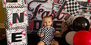 Stetson is registered to the contest to win money with this photo: baby, child, balloons, checkered_pattern, birthday, party, blocks, black, white, red, floor, sitting, smiling, face, head, blue_eyes, background, decorations, racing_flag, onesie