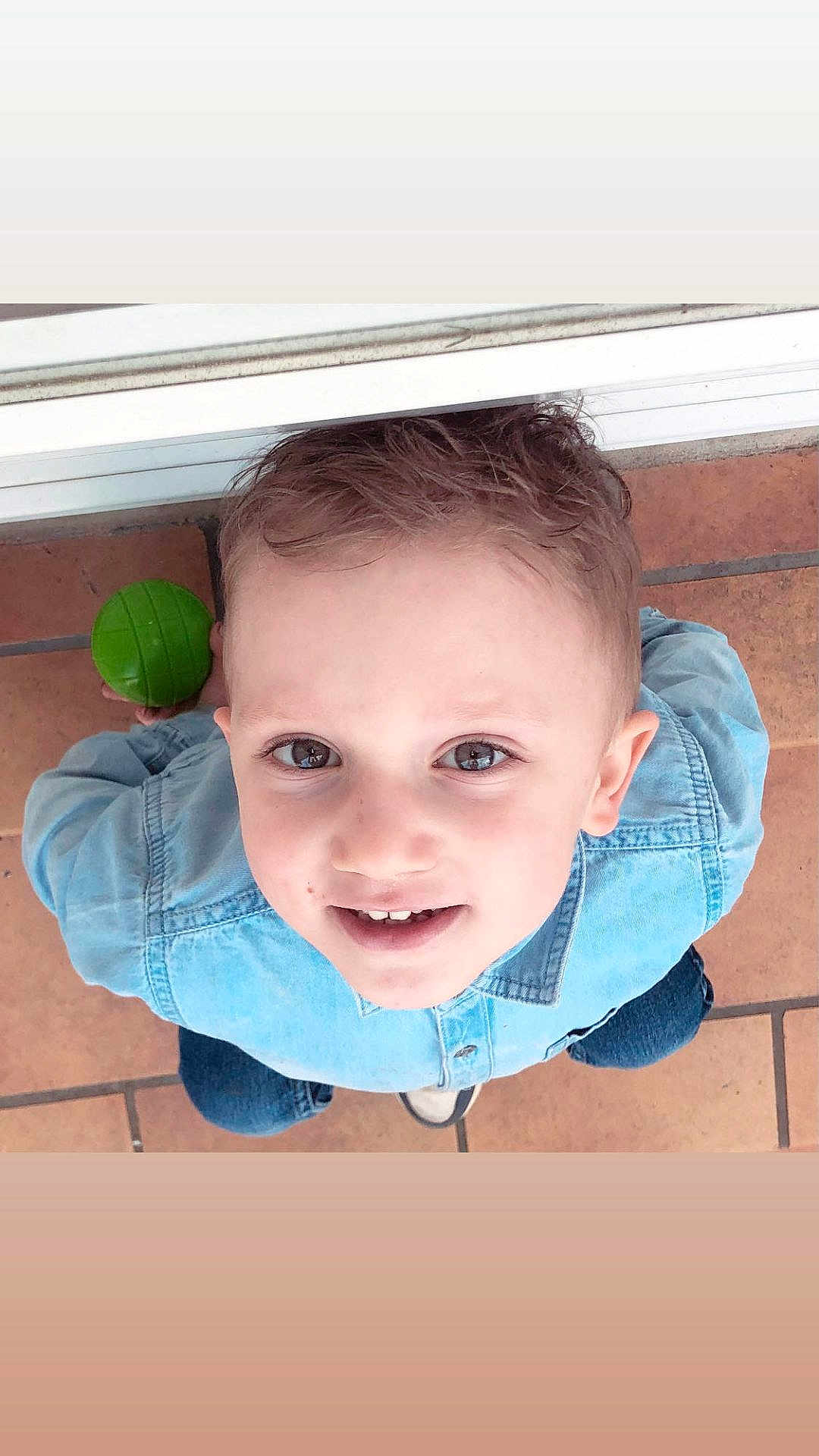 Giullian a rejoint le concours — aidez-le/la à gagner de superbes lots ! child, boy, smiling, face, denim_shirt, green_ball, floor, tile, casual_clothing, short_hair, indoor, playful, looking_up, cute, portrait, happy, person, childhood, young, casual