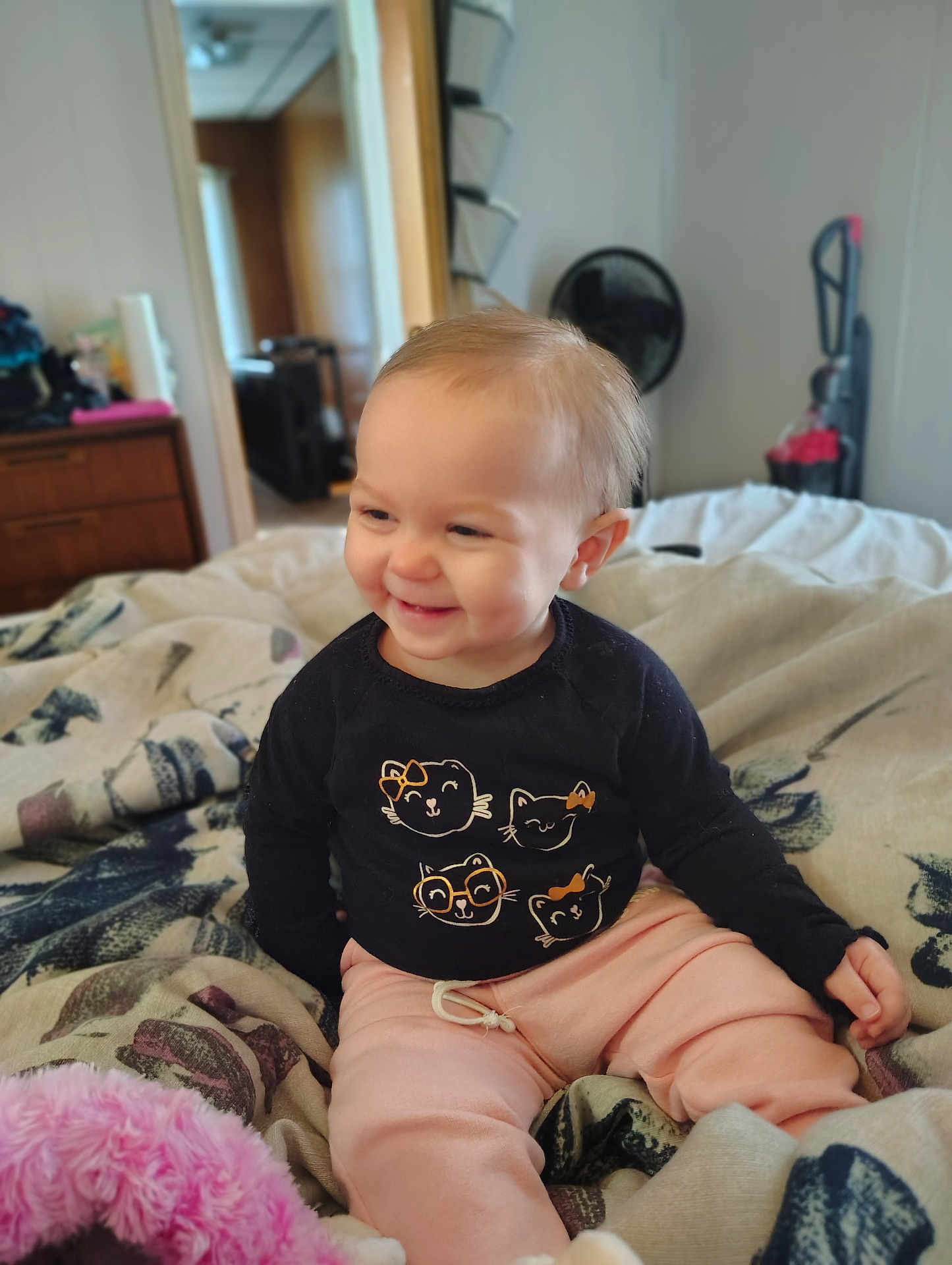 Carlie joined the competition — help win amazing prizes! toddler, child, smiling, bed, blanket, indoor, clothing, person, cute, happy, face, sitting, furniture, pink_pants, black_shirt, cat_graphics, home, soft_toys, cozy, portrait