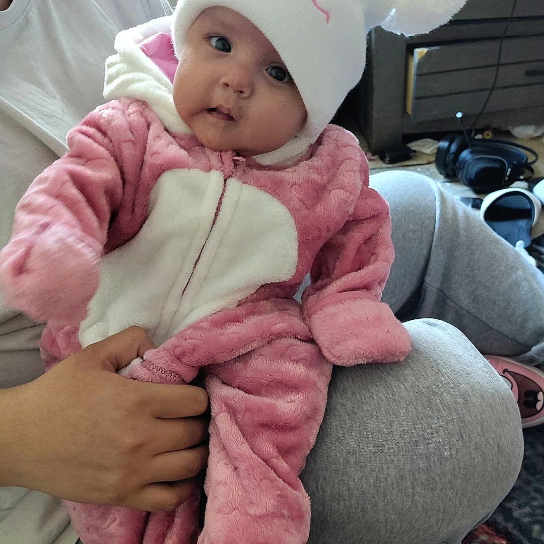 Maylynn Jo joined the competition — help win amazing prizes! baby, casual, child, clothing, cozy, cute, face, floor, furniture, hand, home, indoor, infant, person, pink_outfit, plaid_blanket, pom_poms, smiling, white_hat, window