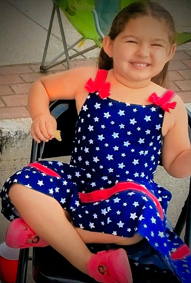 Amara is registered to the contest to win money with this photo: child, smiling, dress, stars, red_bows, pink_shoes, chair, outdoor, snack, happy, person, cute, young, sitting, pavement, footwear, playful, casual, daylight, relaxed