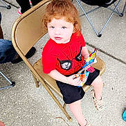 Carl is registered to the contest to win money with this photo: child, toddler, red_hair, spiderman_shirt, black_shorts, folding_chair, outdoor, snack, plastic_shoes, concrete_floor, people, legs, casual_clothing, seated, daylight, bracelet, curly_hair, cute, looking_up, holding