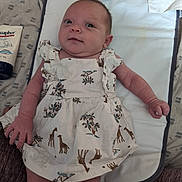 Pennelopea is registered to the contest to win money with this photo: baby, infant, dress, giraffe_pattern, changing_pad, ointment, eucaphor, lying_down, curious_expression, hand, face, skin, clothing, fabric, home, floor, care, newborn, person, cute