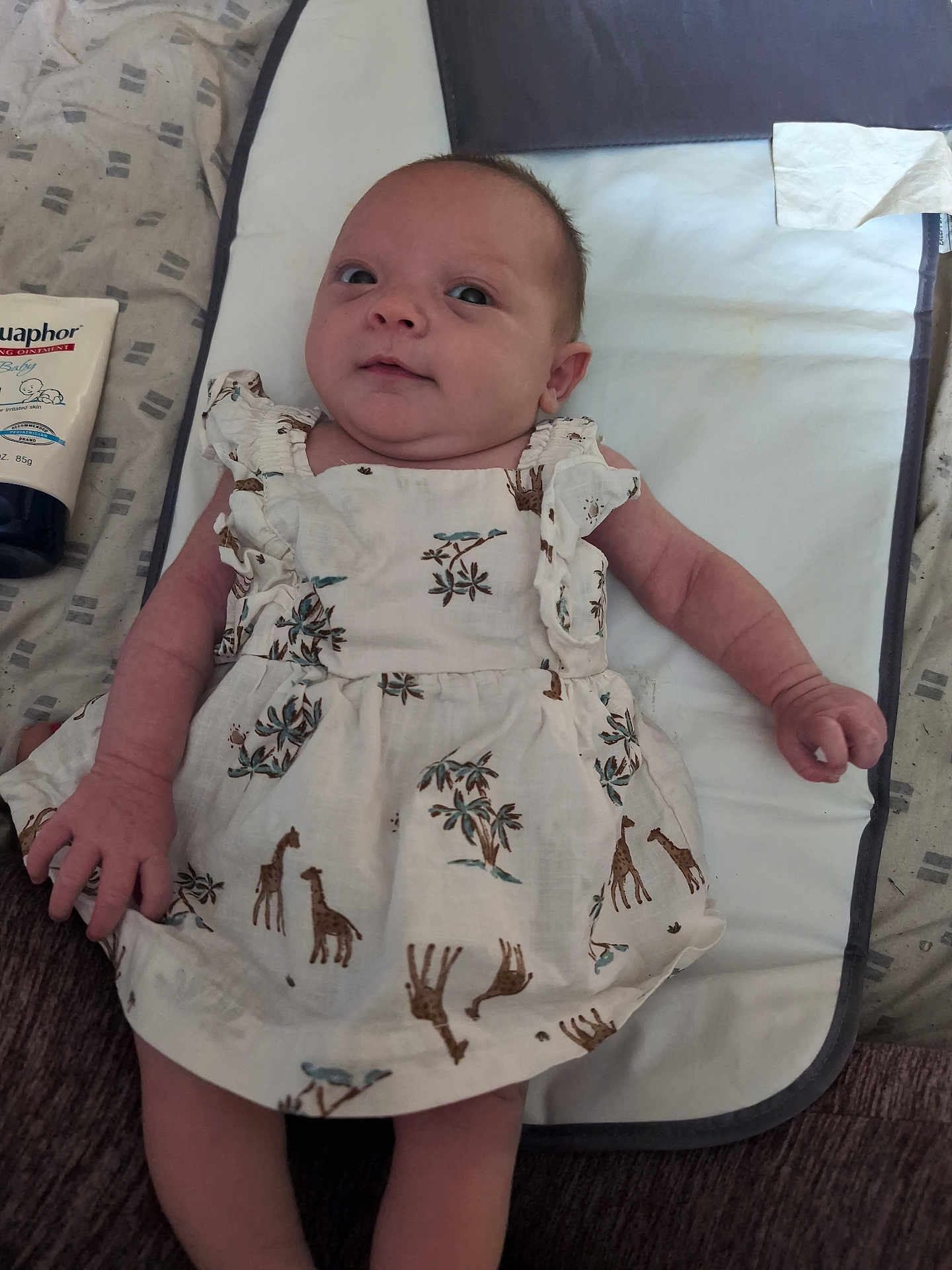 Pennelopea is registered to the contest to win money with this photo: baby, infant, dress, giraffe_pattern, changing_pad, ointment, eucaphor, lying_down, curious_expression, hand, face, skin, clothing, fabric, home, floor, care, newborn, person, cute