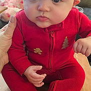 Pennelopea is registered to the contest to win money with this photo: baby, child, infant, red_clothing, santa_hat, holiday_outfit, wooden_surface, hand, face, eyes, pajamas, phone, blurred_background, indoor, person, cute, festive, seated, young_child, cozy