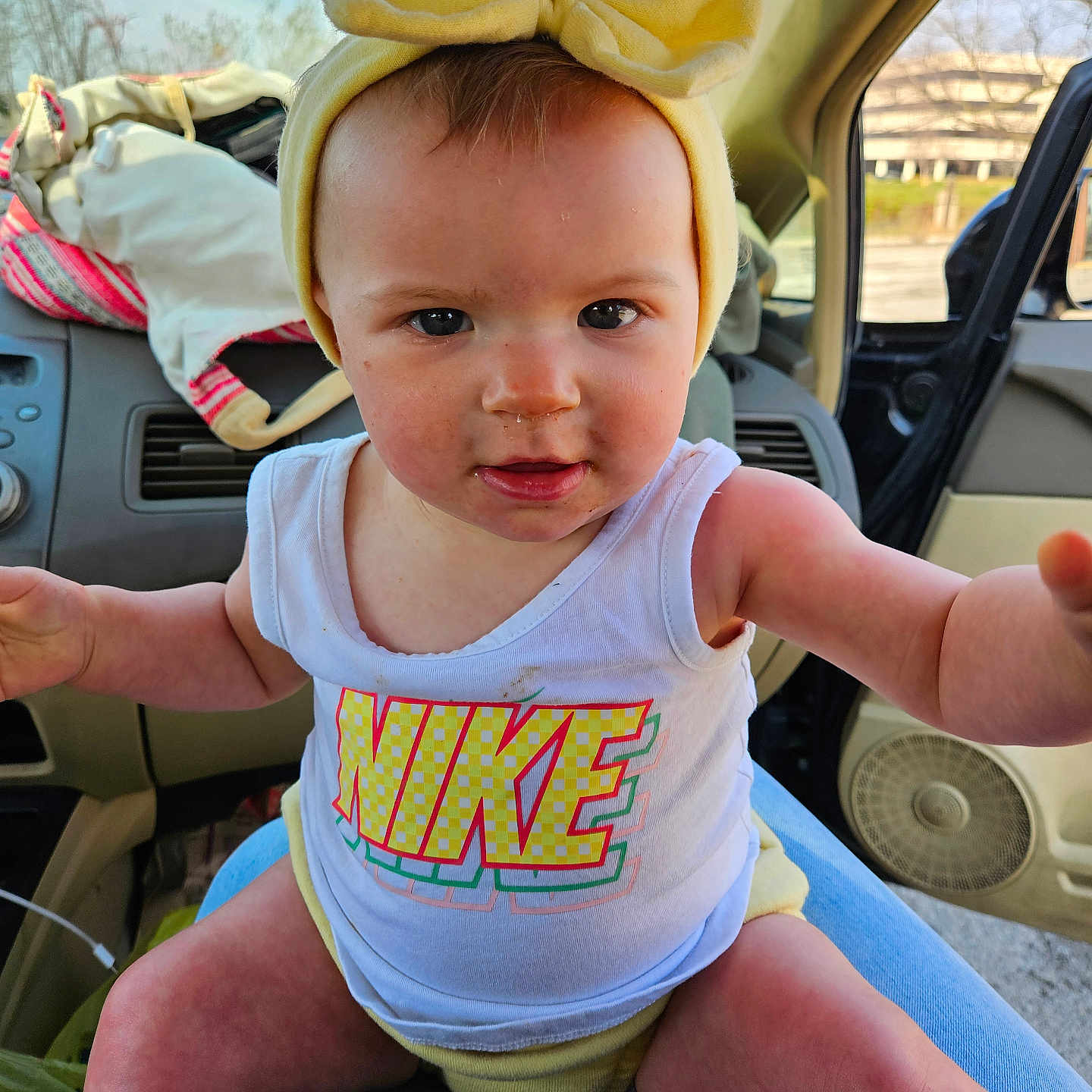 Marlene is registered to the contest to win money with this photo: baby, child, yellow_bow, tank_top, nike_logo, car_interior, person, face, legs, arms, window, seat, clothing, headwear, expression, sunlight, jeans, hand, dashboard, seatbelt